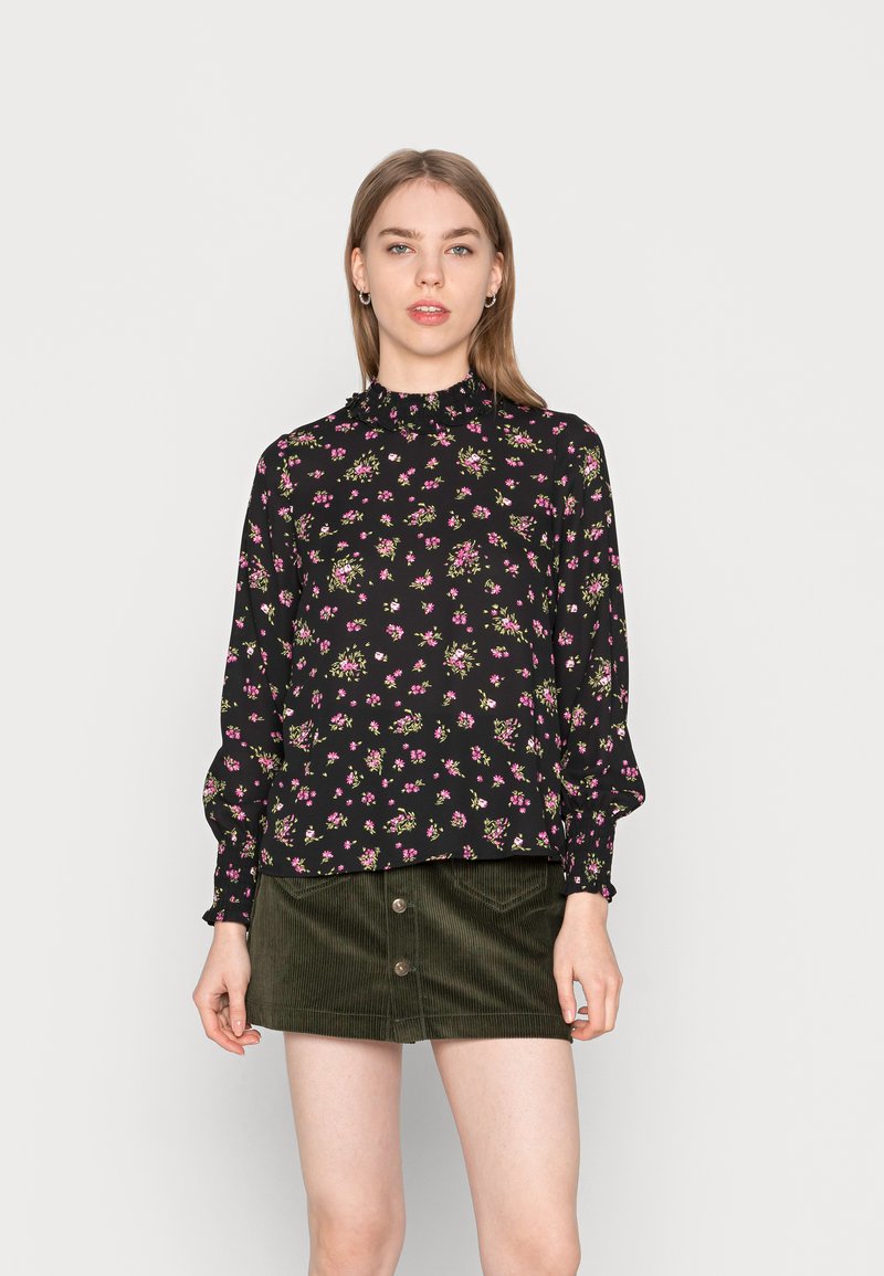 Blusa preta floral com colarinho e mangas com babados, apresentando pequenas flores rosas e verdes. Usada com uma saia de cotim verde escuro com botões.