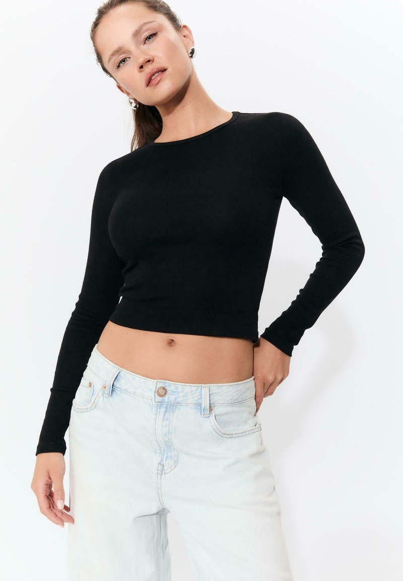 Schwarzes langärmliges Crop-Top aus weichem Stoff mit Rundhalsausschnitt, kombiniert mit hellblauen hochgeschnittenen Jeans mit Knopfverschluss.