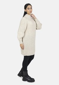 Beige, ribbad klänning med hög krage, oversized ärmar och sidofickor, kombinerad med svarta leggings och snörda ankelstövlar.