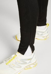 Black athletic pants with a textured finish, featuring side slits at the hem. Paired with white and yellow running shoes.