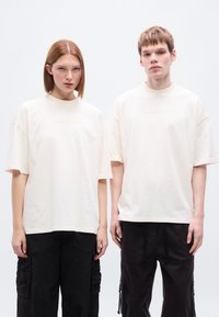 Υπερμεγέθη βαμβακερά t-shirts κρεμ απόχρωσης με ψηλό λαιμόκοψη, κοντά μανίκια και διακριτική κεντητή επιγραφή "BROKE SINCE MILANO" στο μπροστινό μέρος.