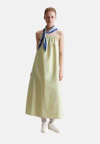Hellgrünes Leinen-Sommerkleid mit elastischem Oberteil, langem, fließendem Rock und einem blauen gemusterten Schal, der um den Hals gebunden ist. Weiße Sneakers.