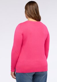 Donna con capelli castani che indossa un maglione a maniche lunghe rosa brillante e jeans blu, mostrata di spalle su uno sfondo chiaro uniforme.