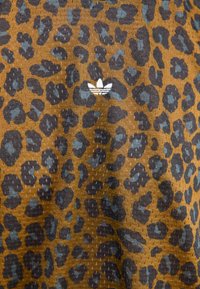 Leopardskinnmönstrad tyg i bruna och mörkblå nyanser, med en broderad vit Adidas-logotyp. Materialet verkar ha en texturerad känsla med subtila mönster.