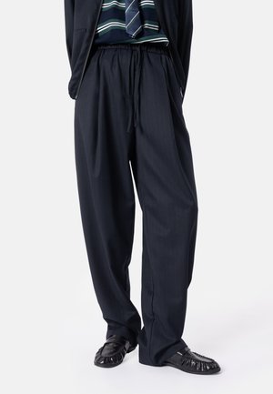 Person trägt weite dunkle Hosen mit Kordelzug in der Taille, schwarze geflochtene Schuhe, ein marineblau gestreiftes Hemd und eine gestreifte Krawatte. Oberkörper nicht sichtbar.