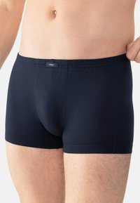 Mand i marineblå tætsiddende boxer shorts, der justerer taljen på en hvid baggrund, viser midtsektionen og øverste lår.