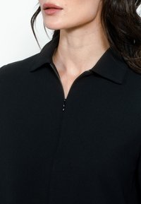 Juodas apvalios apykaklės viršus su trumpu užtrauktuku, pasižymintis lygia, tekstūruota medžiaga ir struktūruota siluetu.