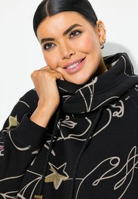 Sweatshirt noir avec un col haut, présentant des motifs artistiques dorés et violets. Le tissu semble doux avec une coupe décontractée. Cordon de serrage inclus.