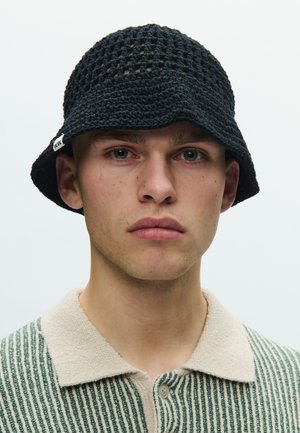 Mladý muž so svetlou pokožkou, ktorý má na hlave čierny háčkovaný klobúk typu bucket a na sebe zeleno-béžové pruhované pletené polo tričko, pozerá sa vpred s neutrálnym výrazom.