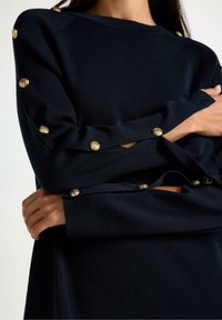 Top in maglia blu navy con maniche lunghe, impreziosito da bottoni dorati lungo le braccia e una vestibilità rilassata. Design pulito con tessuto testurizzato.