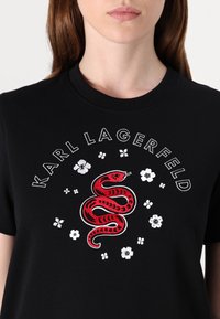 Juodas medvilnės marškinėliai su balta tekstu "KARL LAGERFELD" ir raudonu siuvinėtu gyvate aplink baltų gėlių motyvais.