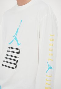 Maglietta a maniche lunghe bianca con logo Jumpman blu e testo grafico nero sul davanti, con scritte stilizzate gialle lungo la manica.