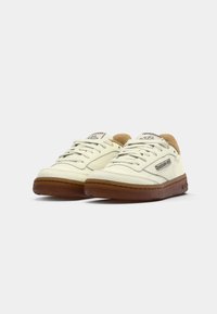Pár krémových tenisiek Reebok s hnedými podrážkami a šnúrkami, vystavených pod uhlom na bielom pozadí.