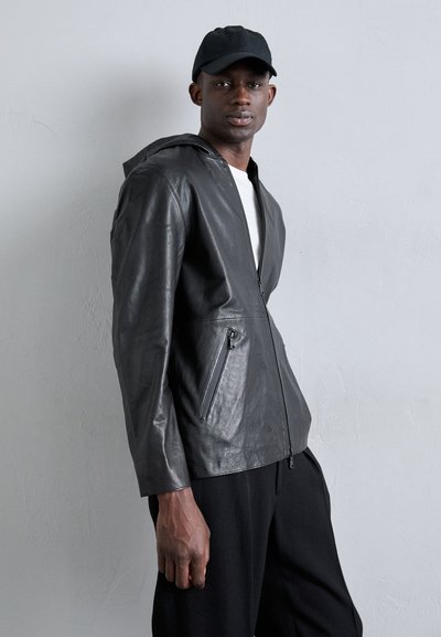 Casaco de pele preta com fecho éclair e capuz, com dois bolsos frontais com fecho éclair e uma textura suave. O modelo veste uma camisa branca por baixo.