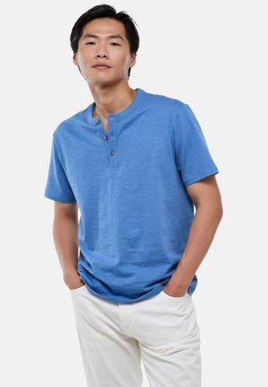 Homme portant un t-shirt henley bleu à manches courtes avec trois boutons et un pantalon blanc, debout les mains dans les poches devant un fond uni.