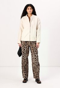 Creamfarbene Fleecejacke, weit geschnittene Leopardenmuster-Hosen, schwarze Tasche und klobige Stiefel. Die Jacke hat einen hohen Kragen und einen Druckknopfverschluss.