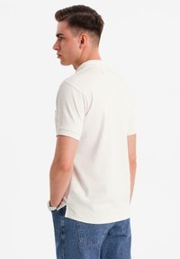 Světle béžové polo tričko s krátkým rukávem, žebrovaným límcem a bočními rozparky, v kombinaci s modrými džínovými kalhotami. Hladká textura látky.