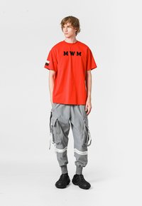 Camiseta de algodón roja con bordado "MWM", pantalones cargo grises con cremalleras y bolsillos de parche, y zapatos negros texturizados.