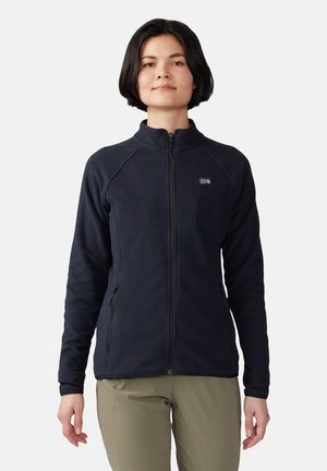 Frau mit kurzen schwarzen Haaren, die eine schwarze Fleecejacke mit durchgehendem Reißverschluss und beige Hosen trägt, steht vor einem schlichten hellen Hintergrund.