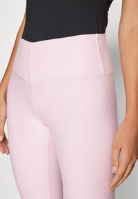 Világos rózsaszín leggings sima textúrával, magas derékkal és lapos varrásokkal a tartósság és kényelem érdekében. Közeli kép a derék és a csípő területéről.