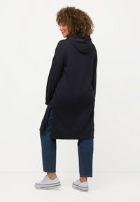 Felpa lunga blu navy con cappuccio, fessure laterali, orlo a coste, indossata sopra jeans blu e sneakers bianche, caratterizzata da una texture morbida e un fit rilassato.