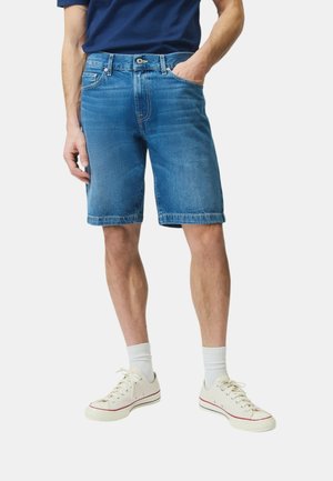 Uomo che indossa pantaloncini di jeans blu di media lunghezza, calzini bianchi e scarpe da tela bianche, in piedi con una mano in tasca.