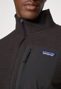 Zwarte fleece jas met een ritssluiting en een gestructureerd patroon. Voorzien van een contrasterende stoffen borstzak met rits en het Patagonia-logo.