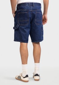 Mężczyzna noszący ciemnoniebieskie szorty dżinsowe z wieloma kieszeniami, białe skarpetki i białe trampki z czarnymi paskami, stojący na jednolitym tle.