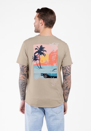 Hombre con brazos tatuados que lleva una camiseta beige con un gráfico de un atardecer colorido, palmeras y un coche vintage en la parte posterior.