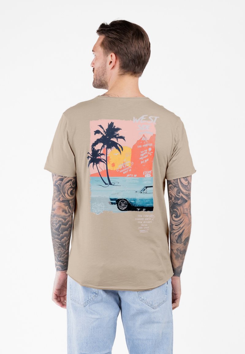 Hombre con brazos tatuados que lleva una camiseta beige con un gráfico de un atardecer colorido, palmeras y un coche vintage en la parte posterior.
