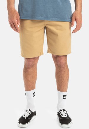 Pantaloni corti beige con taglio dritto, realizzati in cotone. Indossati con sneaker nere e calzini bianchi con un logo. Outfit casual, dettagli minimi.