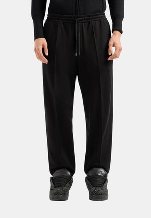 Pantaloni neri con una vestibilità rilassata, dotati di una vita con coulisse, una parte anteriore plissettata e un tessuto liscio. Abbinati a sneaker nere testurizzate.