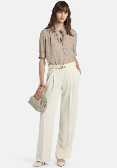 Bež bluza z belimi pikami s kratkimi rokavi, krem hlače z visokim pasom, zlati pas in teksturirana siva torbica. Špičasti bež čevlji.