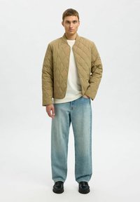 Beżowa pikowana kurtka z zapięciem na napy, w połączeniu z jasnoniebieskimi spodniami o szerokich nogawkach i czarnymi butami, charakteryzująca się okrągłym dekoltem i wzorem w diamenty.