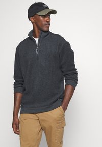 Dunkelgrauer Fleece-Pullover mit Reißverschluss, gerippten Bündchen und Saum, kombiniert mit beigefarbenen Cargo-Hosen. Die Person trägt eine schwarze und olivgrüne Kappe.