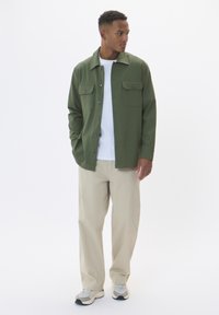 Veste verte à boutons avec deux poches poitrine, portée sur un t-shirt blanc. Assortie à un pantalon beige et des baskets gris clair.