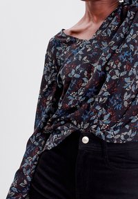 Blouse noire à motifs floraux avec des feuilles bleues et rouges, tissu drapé, large encolure et manches longues. Associée à un jean foncé avec un bouton en argent.
