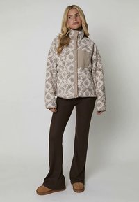 Giacca in pile beige con motivo, collo alto e tasca sul petto, abbinata a pantaloni marroni a zampa e pantofole marrone chiaro.