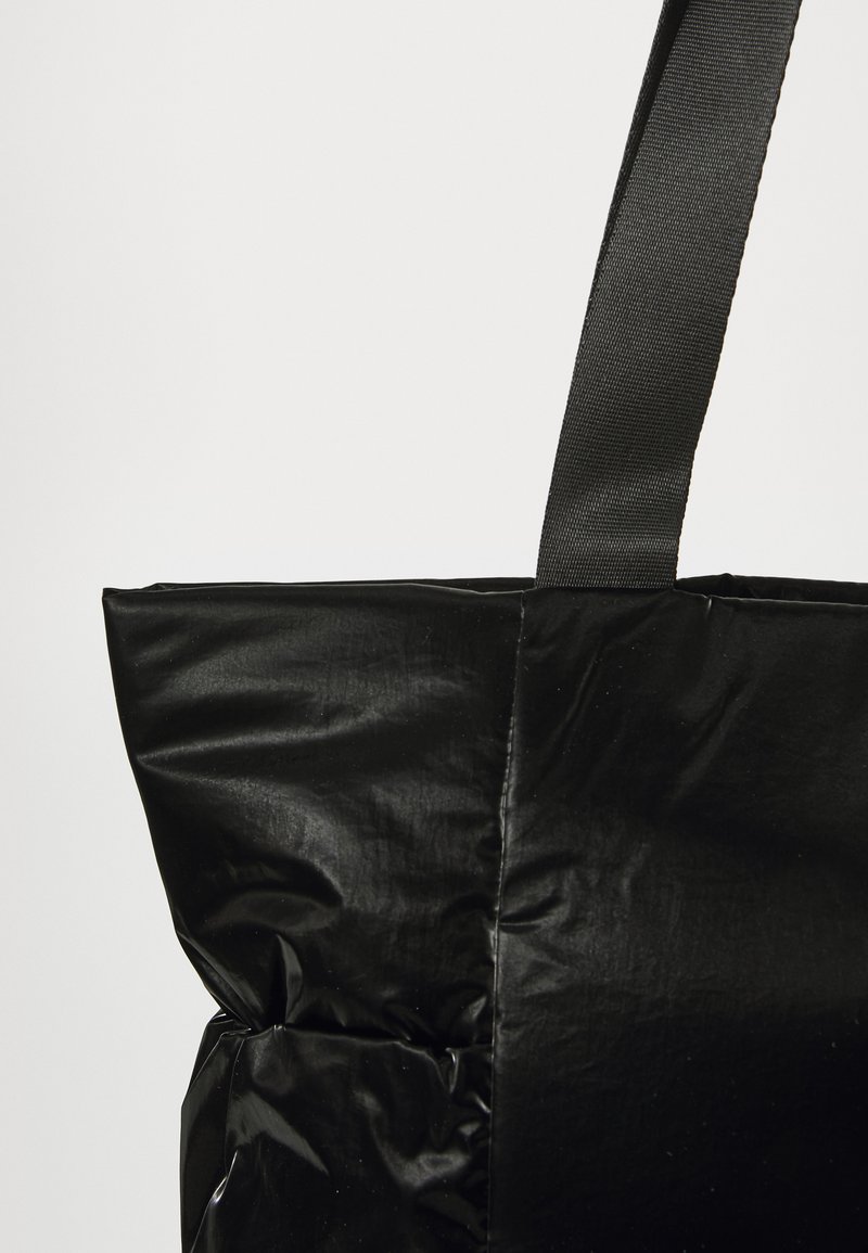 Bolso tote negro con una textura crujiente y brillante. Presenta una correa de tejido negro y un diseño estructurado y acolchado. Sin hardware visible.