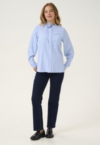 Camicia a righe blu chiaro e bianca con colletto staccabile decorato da una rosa, abbinata a jeans in denim scuri e scarpe nere.