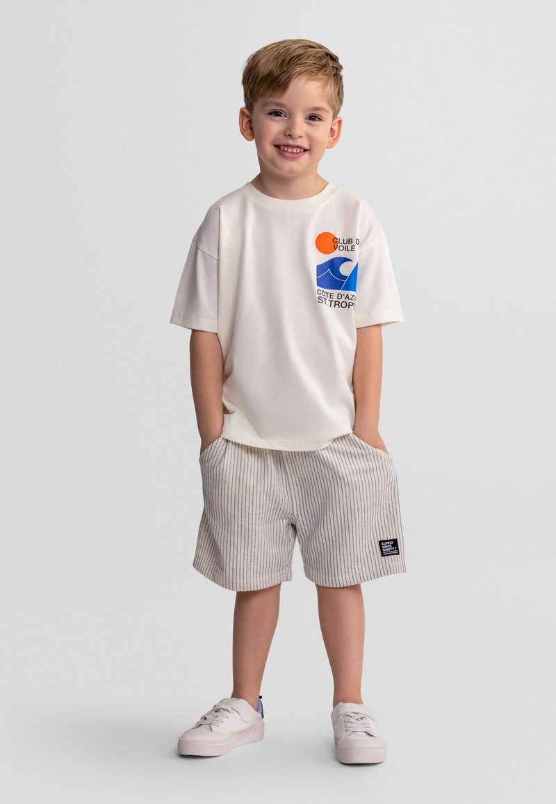 Smiling young boy stands with hands in pockets, wearing white graphic t-shirt, striped shorts, and white sneakers on plain background.