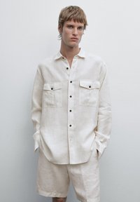 Linen shirt in off-white with a relaxed fit, front buttons, two chest pockets, and contrasting brown buttons. Worn with matching shorts.