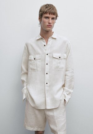 Camicia di lino off-white con vestibilità rilassata, bottoni frontali, due tasche sul petto e bottoni marroni a contrasto. Indossata con pantaloni corti abbinati.