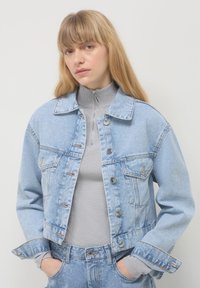 Giacca di jeans azzurro chiaro con un design corto, bottoni frontali in evidenza, due tasche sul petto e cuciture a contrasto. Indossata sopra un dolcevita grigio.