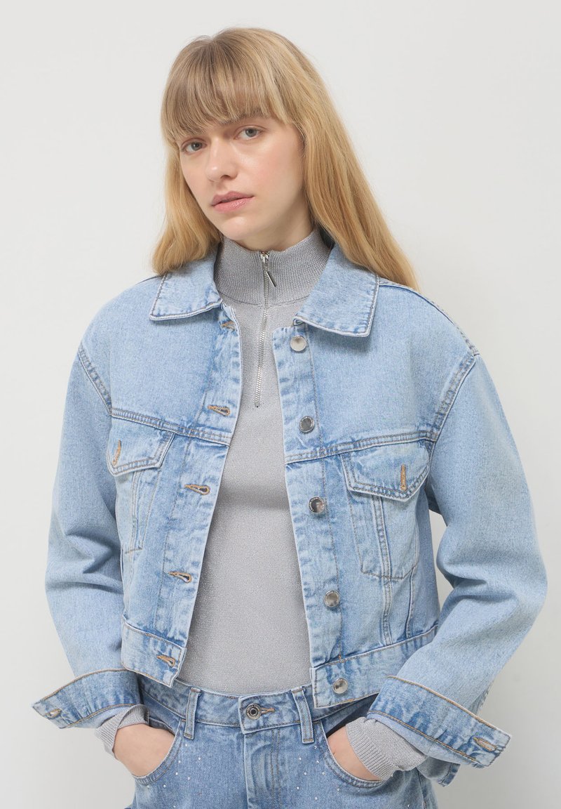 Giacca di jeans azzurro chiaro con un design corto, bottoni frontali in evidenza, due tasche sul petto e cuciture a contrasto. Indossata sopra un dolcevita grigio.