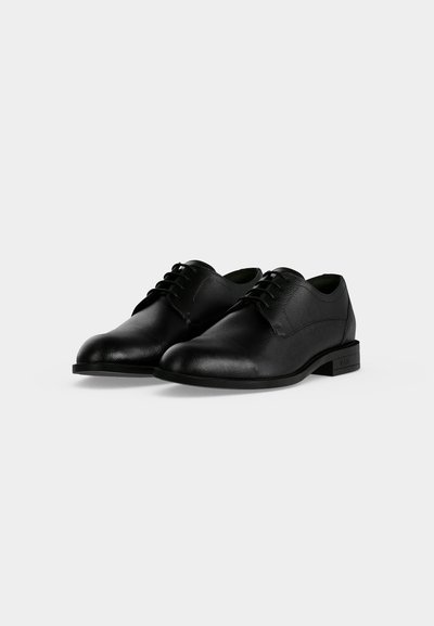 Paire de chaussures habillées en cuir noir à lacets avec petits talons sur un fond gris clair uni.