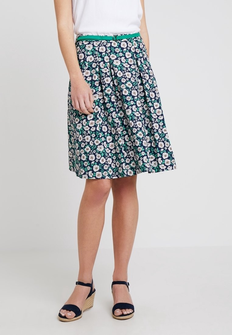 Femme portant une jupe bleu marine longueur genoux avec imprimé floral blanc et vert, une ceinture verte, un top blanc et des sandales compensées bleu marine.