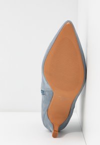 Une chaussure en daim bleu clair à bout pointu avec une semelle en caoutchouc beige, positionnée debout contre un mur blanc en angle.