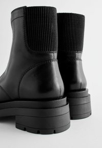Svarta läderankelboots med ribbade elastiska paneler, platta gummisulor och slät yta. Har en tjock klack med texturerad detalj.