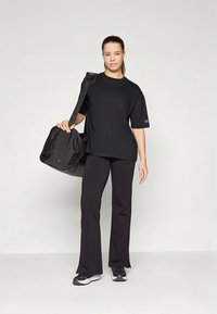 Zwart oversized T-shirt met korte mouwen, gecombineerd met zwarte uitlopende broeken. Een zwarte sporttas hangt over één schouder.